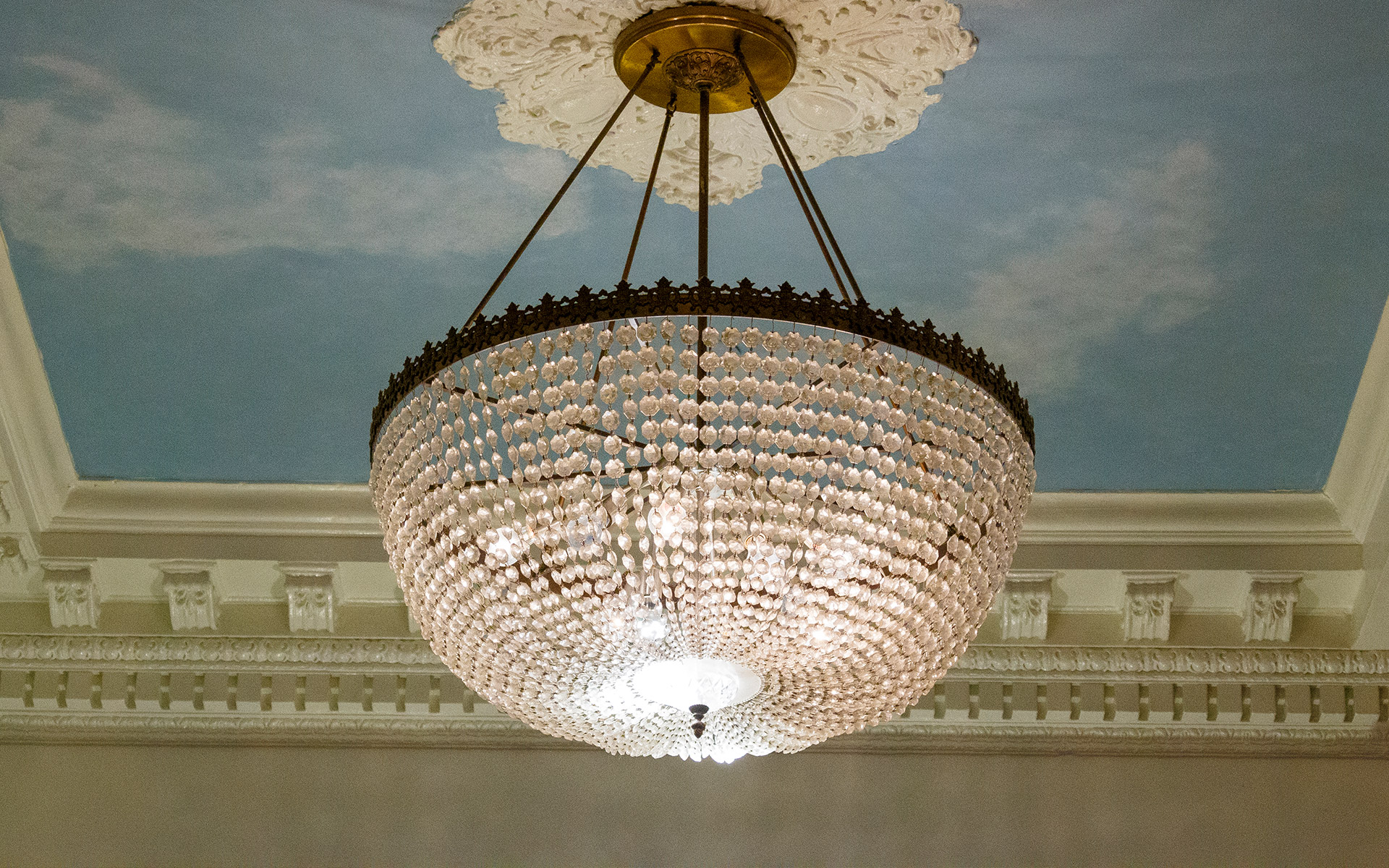 a large chandelier hanging from a ceiling with a cloudy sky mural