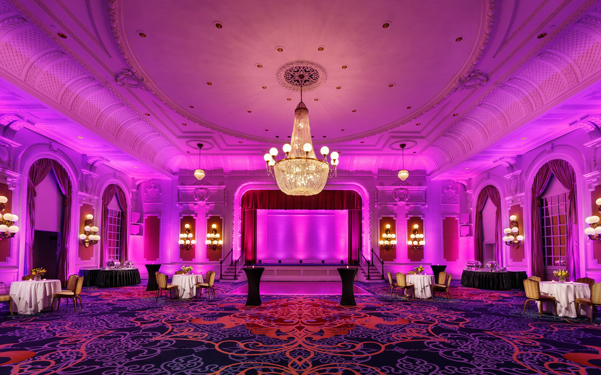 A large, ornate meeting room with purple lighting, a stage, and tables and chairs arranged for an event.